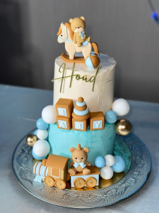Photo de galerie - Gâteau d’anniversaire personnalisé à deux étages, dans les tons bleu, blanc et beige, avec décorations sur le thème bébé.  Réalisation sur mesure selon le thème et les couleurs souhaités.