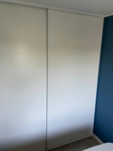 Photo de galerie - Montage de portes coulissantes pour armoire encastrée : ajustement précis des panneaux blancs, pose des rails haut et bas, mise à niveau et alignement parfait sur cloison existante. Espace rangement intégré et fonctionnel.
