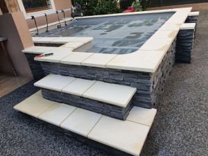 Photo de galerie - Piscine surélevée moderne en pierre reconstituée grise avec margelles claires et escalier maçonné assorti, alliant solidité et finition soignée.

