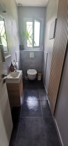 Photo de galerie - WC indépendant élégant : mur en béton ciré gris, WC suspendu, petit lave-mains sur meuble bois, carrelage noir au sol, claustra bois sur le côté et fenêtre avec plante verte.