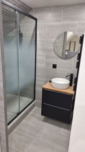 Photo de galerie - Salle de bain rénovée : carrelage moderne, douche vitrée avec profil noir, meuble vasque design avec robinet noir et miroir rond.
