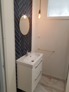 Photo de galerie - Rénovation d'une salle d'eau de studio à Bayonne. Pose d'une crédence en faïence disposée en chevrons et d'un sol effet marbre. Installation d'un meuble vasque suspendu, d'un miroir circulaire et d'accessoires de finition dorés.