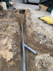 Photo de galerie - Réalisation d’une tranchée pour raccordement au tout-à-l’égout avec pose de canalisation PVC, lit de sable et préparation avant remblaiement.
