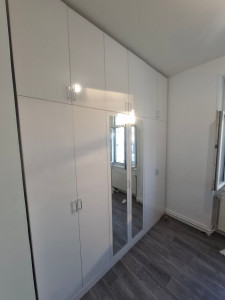 Photo de galerie - Chambre moderne avec une grande armoire blanche brillante sur mesure, installée jusqu’au plafond. L’armoire possède plusieurs portes avec poignées argentées et deux miroirs au centre. Sol effet parquet gris et murs blancs, ambiance propre et élégante.