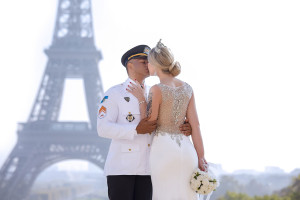 Photo de galerie - Moment romantique d’un couple en uniforme et robe de mariée scintillante, s’embrassant avec la Tour Eiffel en arrière-plan. Une scène élégante et symbolique, pleine de tendresse et d’amour.