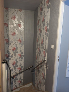 Photo de galerie - Constat de l’état des murs dans la cage d’escalier avant travaux : papier peint arraché, surfaces irrégulières, défauts de planéité et zones de mur détériorées nécessitant une reprise complète avant pose de la nouvelle tapisserie.