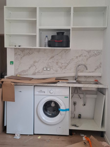 Photo de galerie - Cuisine en cours d’installation : meubles hauts posés, électroménagers mis en place (frigo, machine à laver), évier en cours de raccordement, avec outils et cartons indiquant une phase de finition
