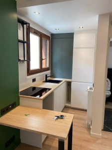 Photo de galerie - A pièce est organisée autour d’une kitchenette en angle, avec des meubles bas blancs au design minimaliste, associés à un plan de travail effet bois clair qui apporte chaleur et contraste. L’évier noir et la plaque de cuisson encastrée renforcent l’aspect