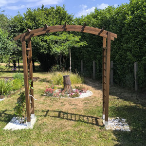 Photo de galerie - Implantation d'une arche décorative, plantage de rosiers grimpant de chaque côté (en cours), puis valorisation d'une souche d'arbre avec plantations  entourées de pierres cimentées et lit de cailloux blancs.