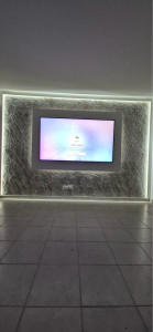 Photo de galerie - Mur TV décoratif avec revêtement effet pierre, encadrement en panneaux MDF et éclairage LED indirect autour de la télévision et sur le pourtour du mur, pour un rendu moderne et épuré