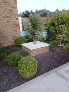 Photo de galerie - Jardinière maçonnée habillée de pierre naturelle et finie par des couvertines en travertin beige. L'olivier et les galets blancs apportent une touche inspirée des jardins méditerranéens, pour un rendu élégant, durable et intemporel.